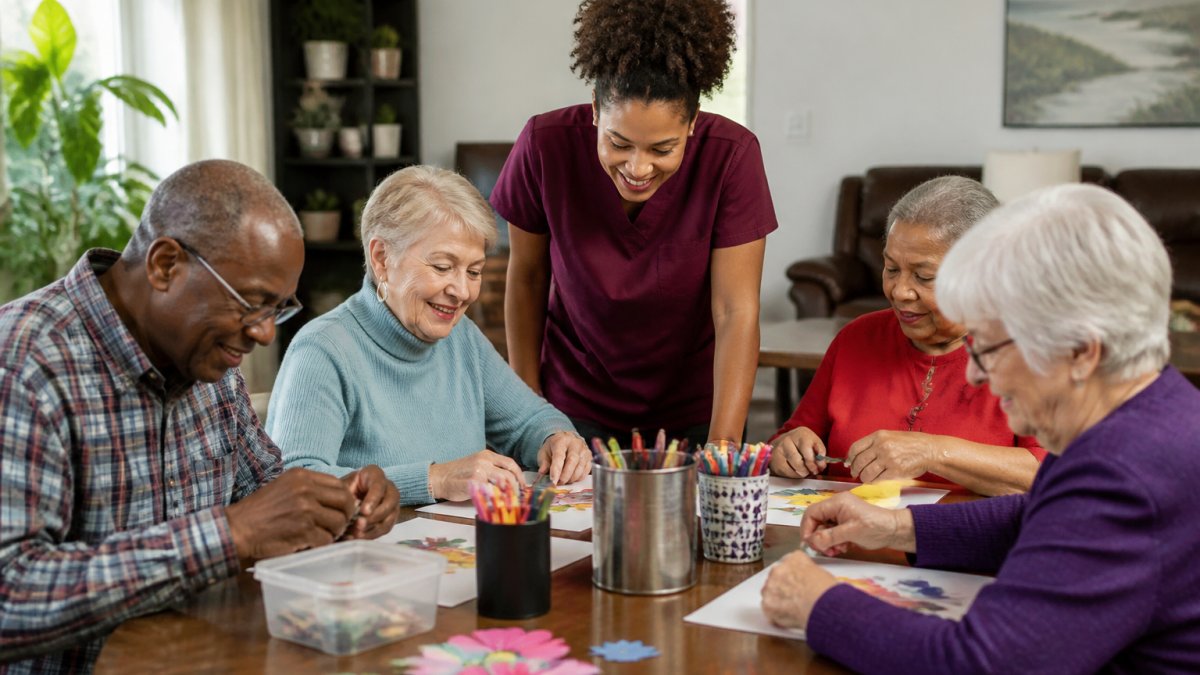 A small group of older adults seated around a table doing a craft activity with a friendly staff member guiding them in a bright, home-like room