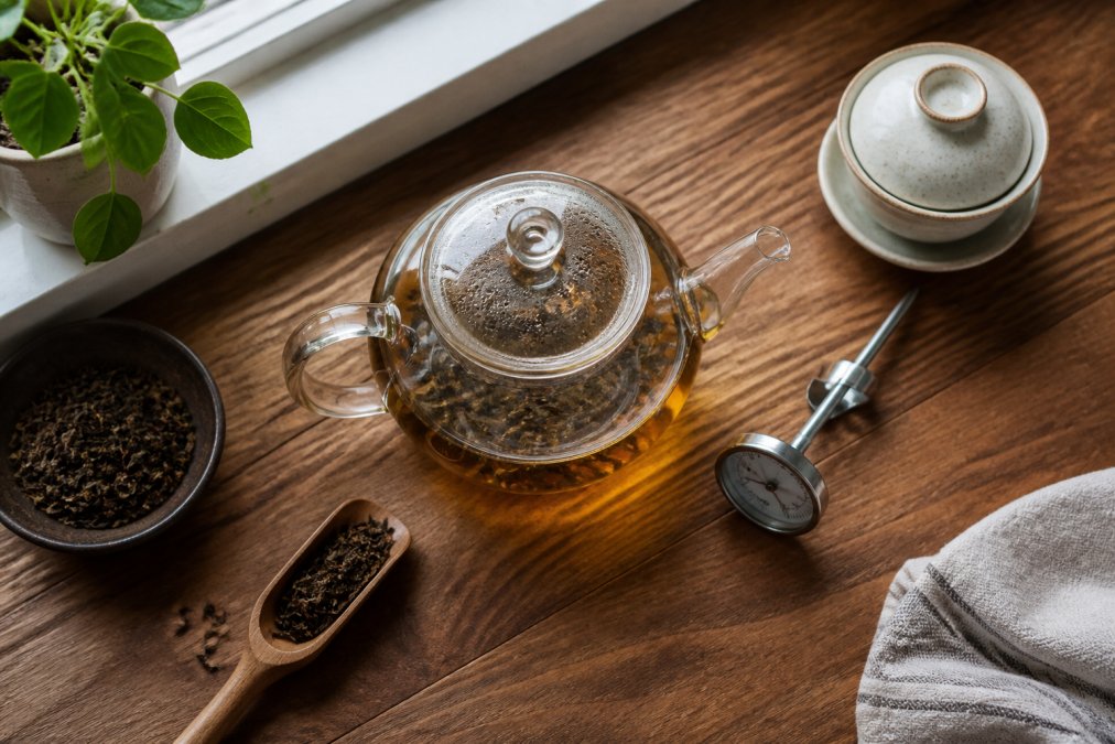 Overhead shot of a glass teapot with loose leaf tea steeping, a small digital thermometer, and a ceramic gaiwan on a wooden kitchen counter with natural light