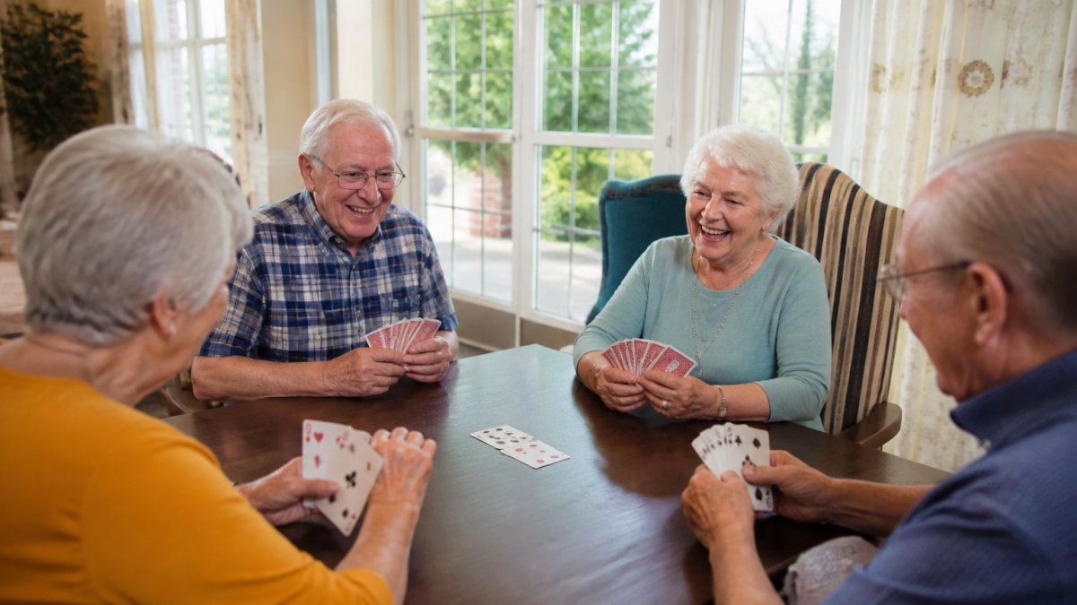 A small group of older adults sitting around a table playing cards and laughing together in a bright, comfortable community room with large windows