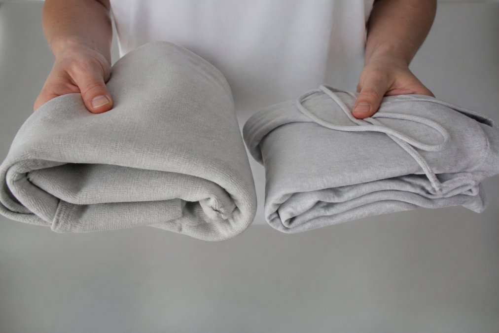 Close-up of hands holding a thick, structured 300GSM cotton hoodie next to a thin fast-fashion hoodie, showing the difference in fabric weight and texture on a neutral background