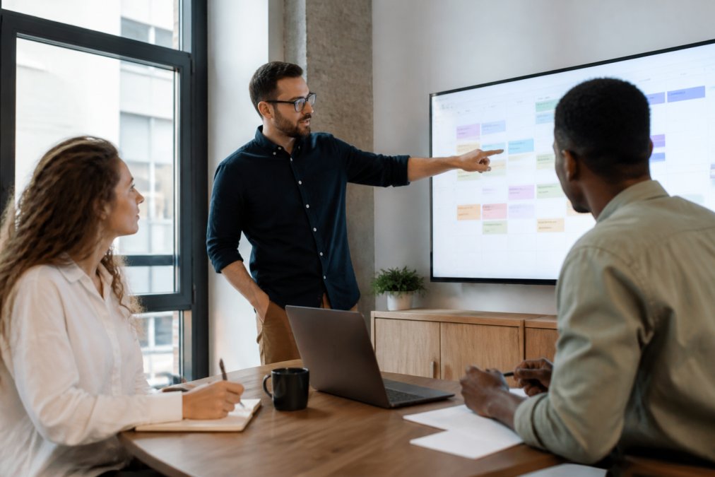 A content team collaborating in a bright modern office, two people reviewing a publishing schedule on a large monitor while a third takes notes