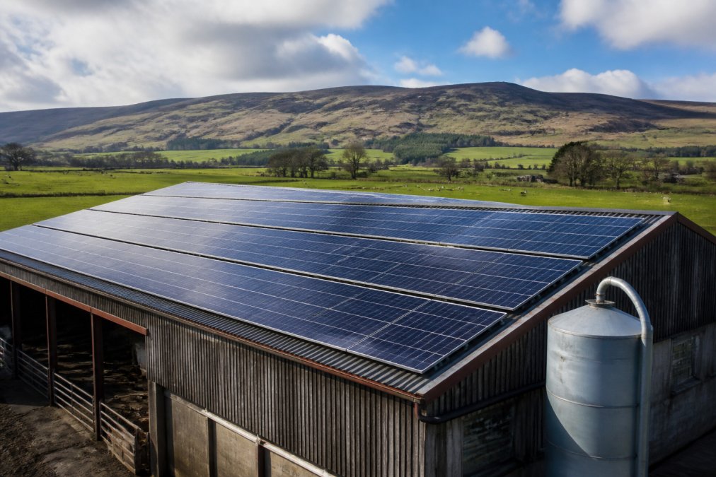 Wide shot of a large agricultural barn roof covered in solar PV panels on a Scottish farm, with green fields and hills in the background on a bright day