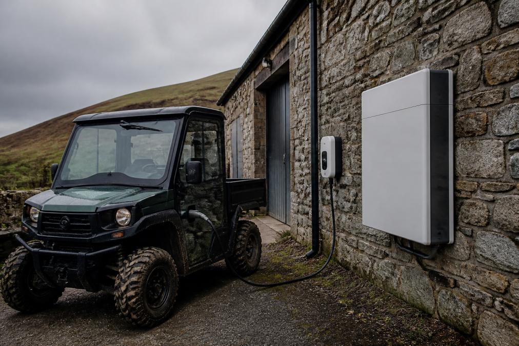 A modern electric farm utility vehicle charging from an outdoor EV charging point beside a rural stone farm building, with a wall-mounted battery storage unit visible and overcast Scottish sky in the background