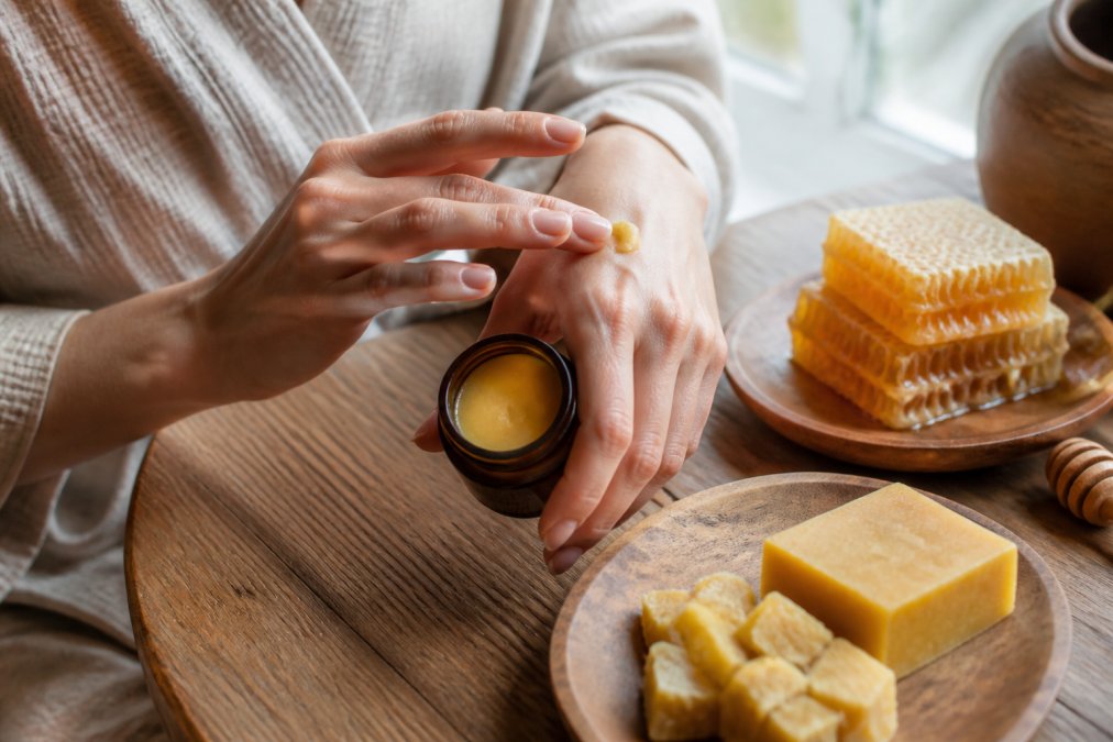 Close-up of a woman's hands applying a golden beeswax balm from a small amber jar, with honeycomb and raw beeswax pieces visible on a wooden surface nearby, warm natural light from a window illuminating the scene