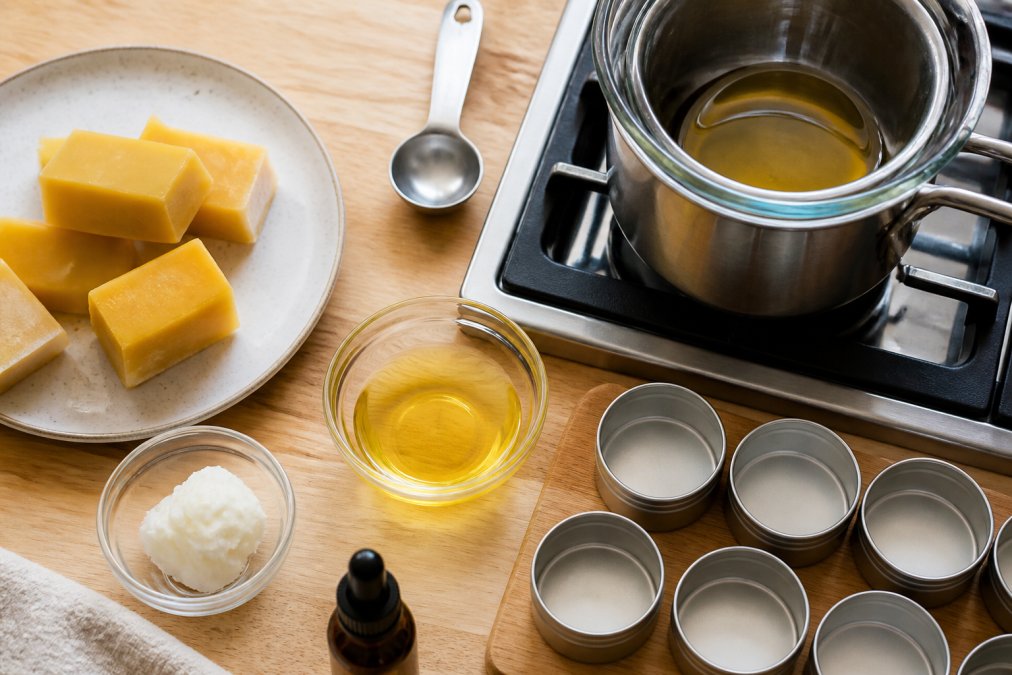 Overhead flat lay of DIY skincare ingredients including small blocks of raw beeswax, a measuring spoon, a double boiler on a stovetop, and small metal tins ready for pouring lip balm or salve, natural light on a light wooden kitchen counter