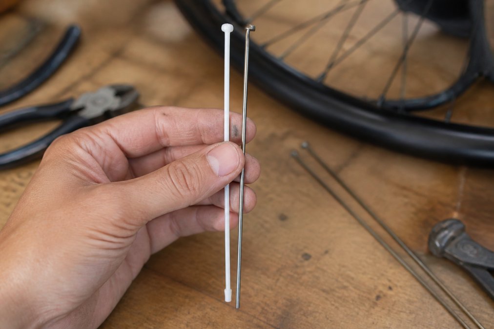 Close-up of a hand holding a single white Berd polymer spoke next to a thin silver steel spoke on a wooden workbench, workshop lighting