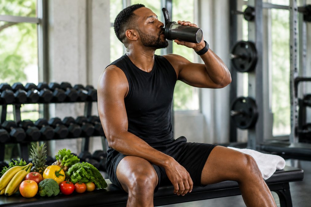 A plant-based athlete in athletic wear drinking from a shaker bottle after a workout in a bright gym, with fresh fruits and vegetables visible nearby on a bench