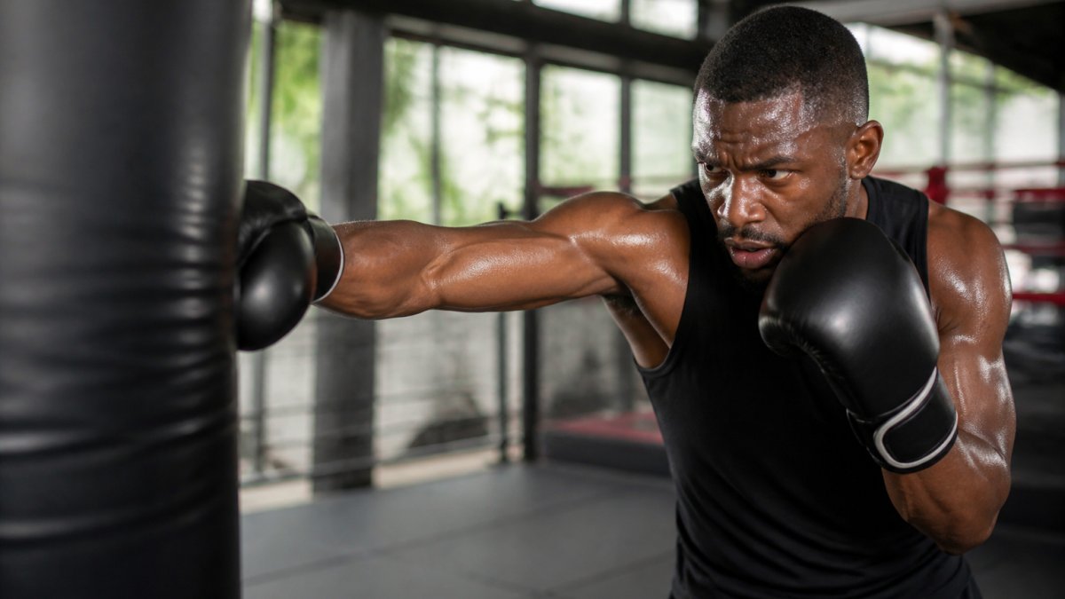 Professional boxer throwing a powerful punch at a heavy bag in a well-lit gym, showing proper form and intensity with sweat visible and focused expression