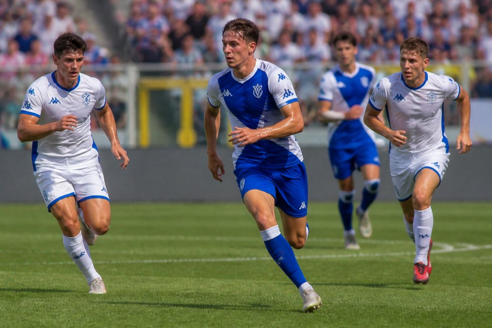 A group of football players in lightweight match kits running across a grass pitch during a competitive match, sweat visible on their faces, bright natural daylight