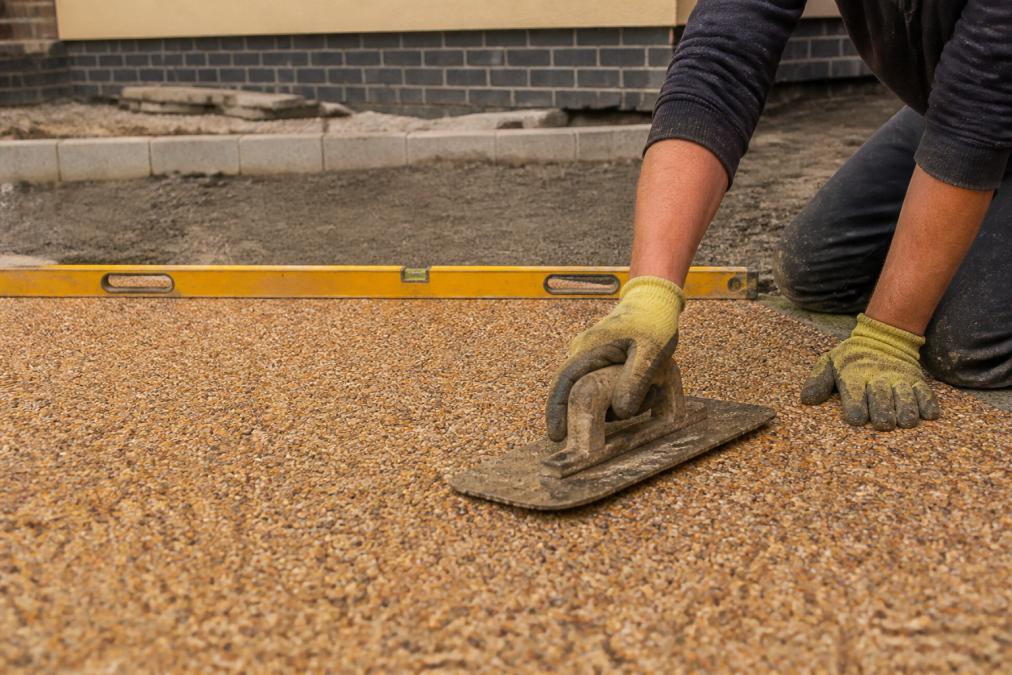A close-up photograph of a contractor's hands smoothing a resin bound surface on a driveway, with a spirit level and compacted sub-base visible in the background