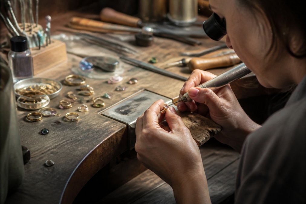 Jeweller's hands at work on a handcrafted piece, with precision tools, coloured gemstones, and precious metals visible on the workbench in warm natural studio lighting