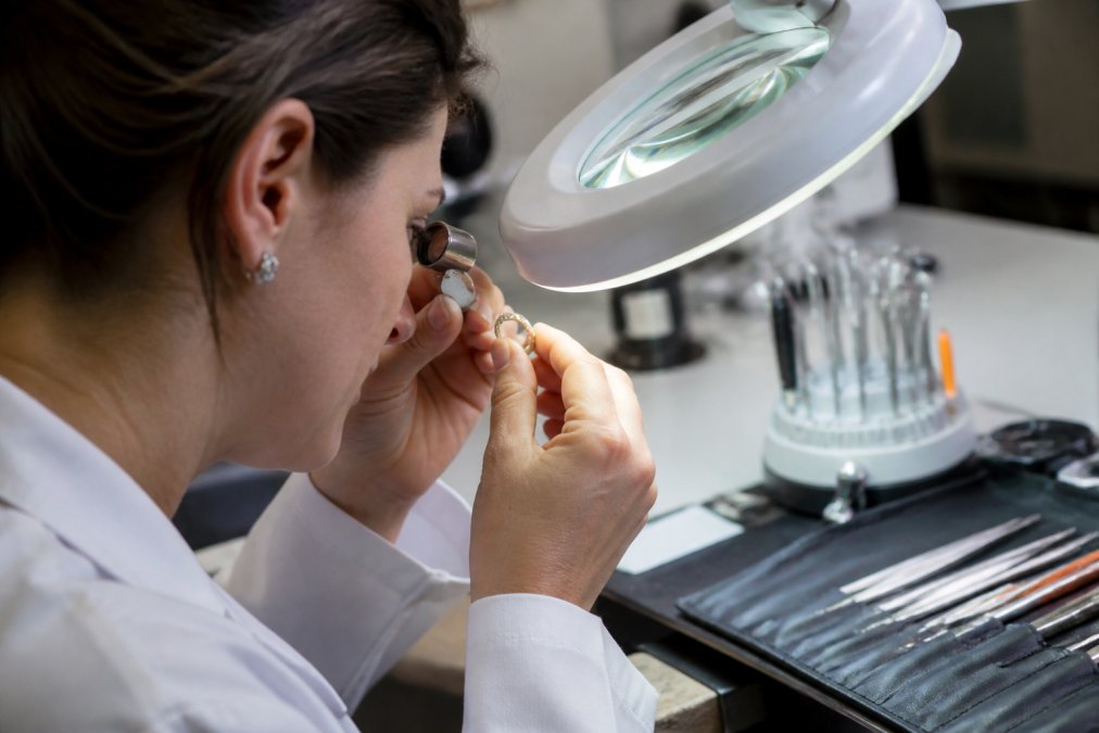 Professional jeweler examining luxury designer ring under magnification lamp, inspecting hallmarks and maker's marks on precious metal band with loupe in bright studio setting
