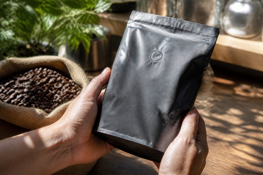 Close-up of hands holding a sealed coffee bag with visible roast date label and one-way valve, sunlight illuminating the rich, glossy surface of freshly roasted beans with their deep brown color and texture