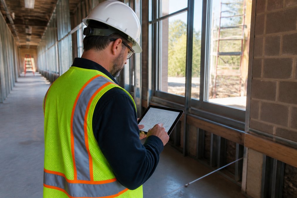 A general contractor in a high-visibility vest reviewing access door specifications on a tablet inside a partially framed commercial corridor, natural light through unfinished window openings