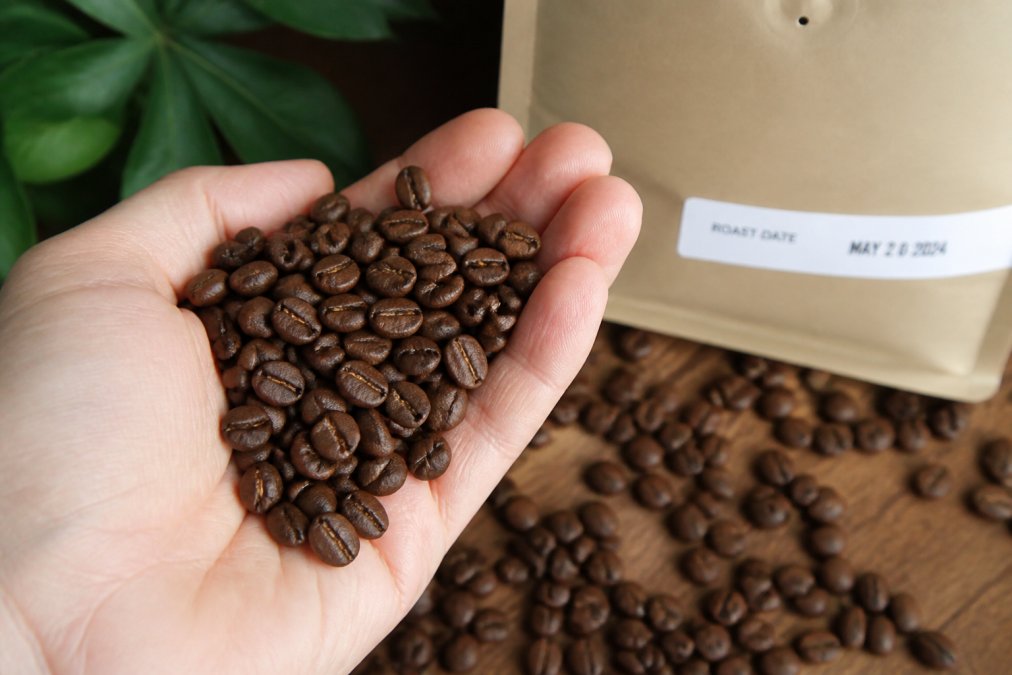 Close-up of freshly roasted coffee beans in a hand showing vibrant brown color and visible bloom, with a roast date label visible on the bag in the background