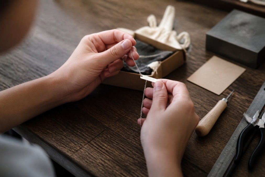 Close-up of hands inspecting the engraved detail on a personalized stainless steel name necklace, with a small maker's card and soft packaging in the background