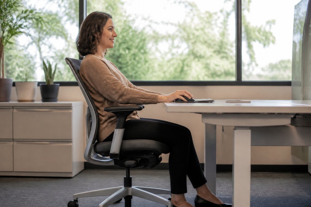 Office professional sitting in an ergonomic Steelcase chair at a desk with proper lumbar support, demonstrating correct posture with arms resting on adjustable armrests, natural window lighting illuminating the workspace