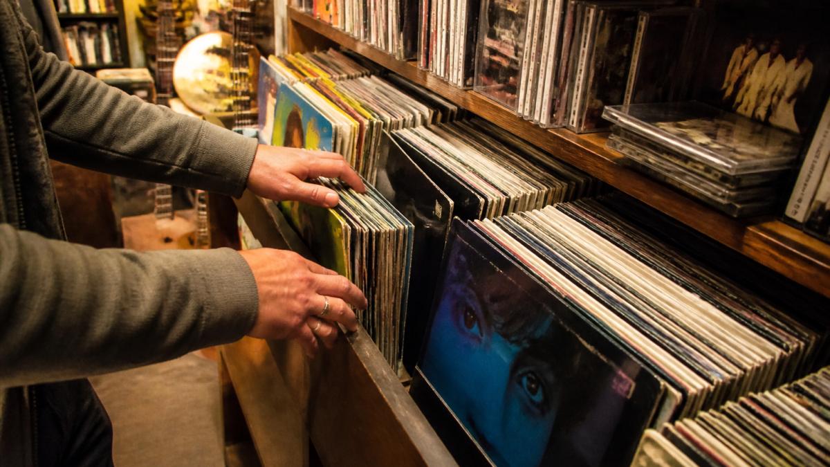 Close-up of hands browsing through vinyl records and CDs on wooden shelves with warm lighting highlighting colorful album artwork and vintage cases