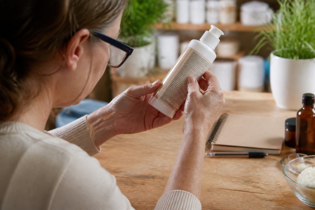 Close-up of hands holding a skincare product bottle, examining the ingredient label carefully with reading glasses
