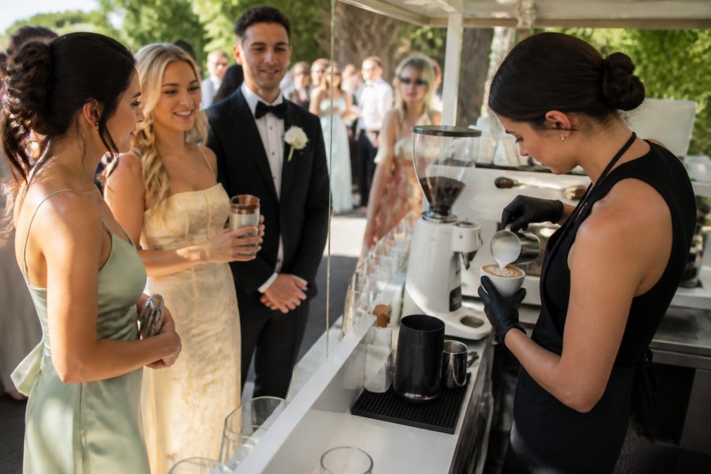 Professional barista preparing specialty lattes with intricate latte art designs for wedding guests gathered around a mobile coffee cart at an outdoor garden venue, with elegantly dressed attendees mingling in soft afternoon sunlight