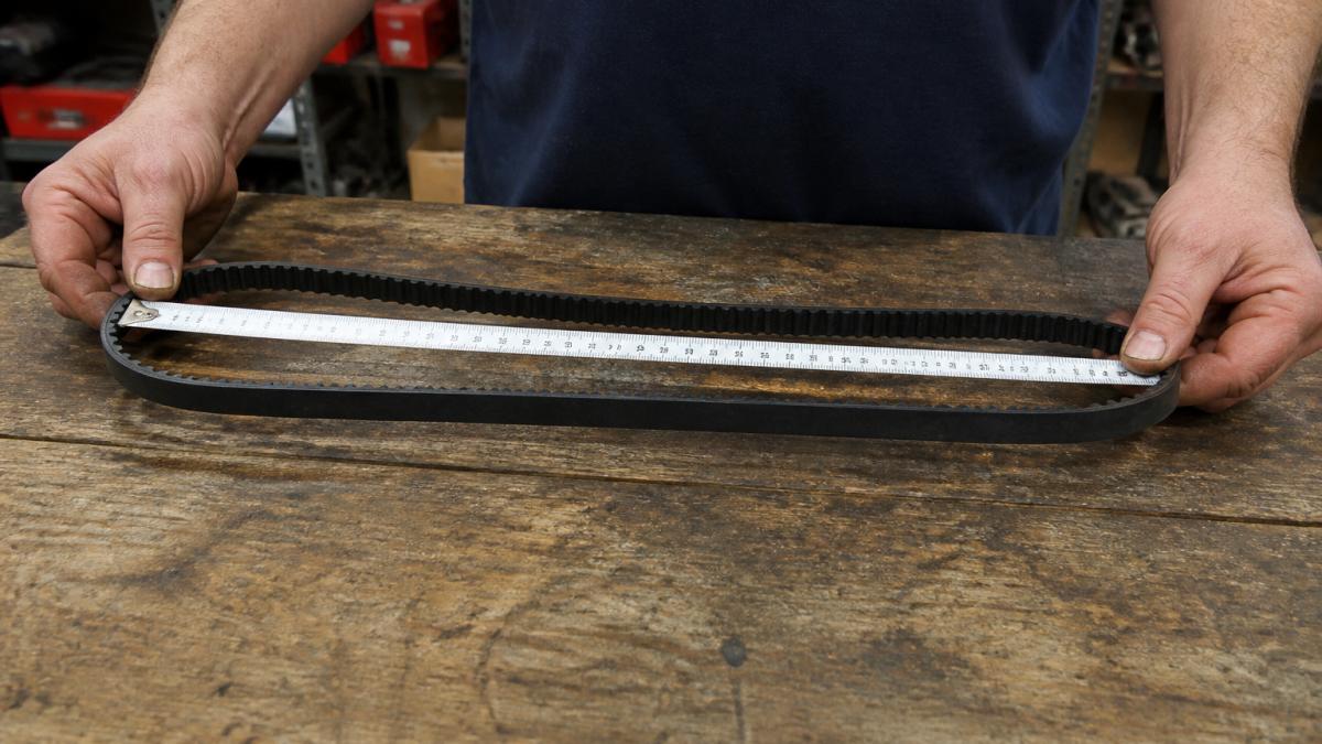 Hands holding a lawn mower belt stretched across a wooden workbench with a fabric measuring tape extended along the belt's length, showing clear measurement markings