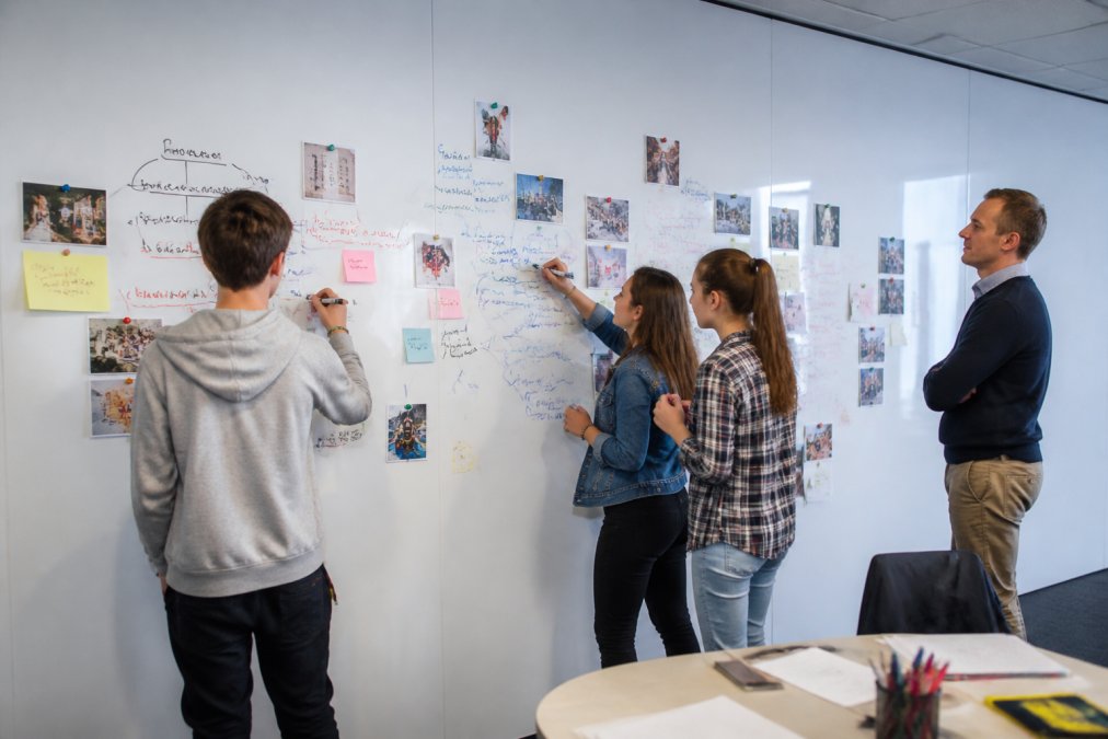 A bright secondary classroom with a floor-to-ceiling magnetic whiteboard wall covered in pupils' handwritten mind maps, sticky notes and photographs, with three teenagers standing and adding to it while a teacher observes
