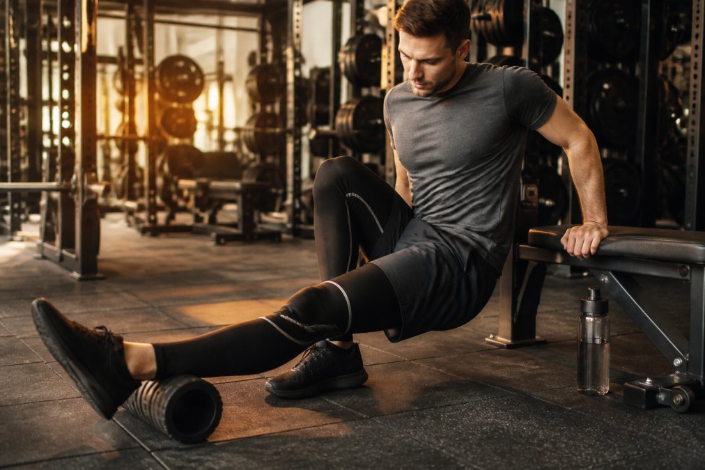 An athlete sitting on a gym bench post-workout, wearing dark compression tights and a knee sleeve, gently stretching one leg forward with a foam roller and water bottle resting beside them under warm gym lighting