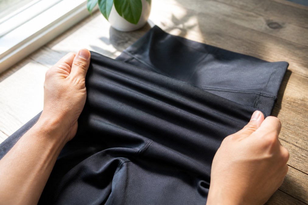 Close-up of a person's hands stretching a high-waisted compression legging fabric between both hands to demonstrate elasticity and texture, photographed under bright natural window light on a wooden surface