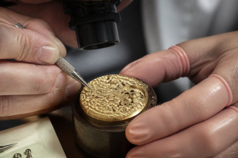 Watchmaker's hands performing perlage finishing on a mechanical movement under magnification, with fine circular patterns visible on the brass plate, professional workshop lighting