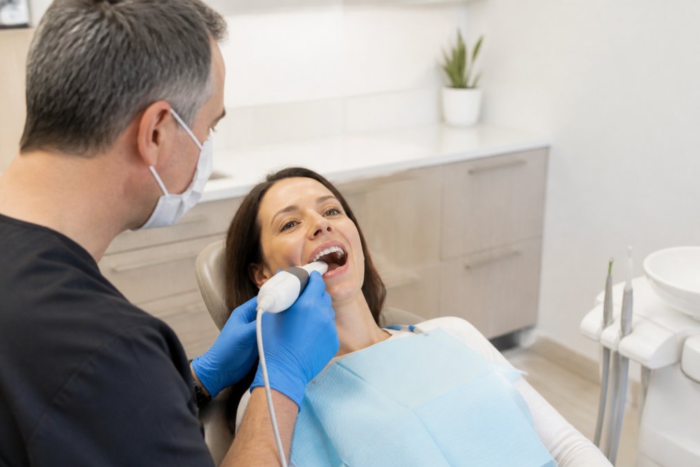 A relaxed female patient sitting in a modern, bright dental chair while a dentist in blue gloves uses a small handheld intraoral scanner wand inside her mouth, with a large monitor displaying a detailed 3D model of teeth beside them