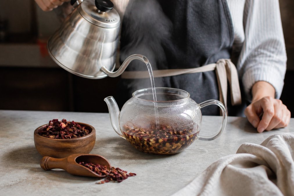 Close-up of hands pouring steaming water from a kettle into a glass teapot filled with loose leaf herbal tea, with a small bowl of dried hibiscus flowers and a wooden scoop nearby on a kitchen counter