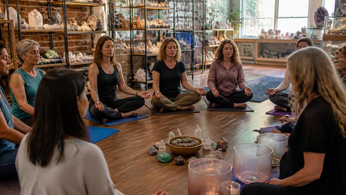 A small group of adults seated in a circle on yoga mats in a softly lit metaphysical shop, eyes closed, with a facilitator gently striking a crystal singing bowl nearby