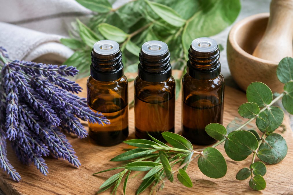 Close-up of amber glass bottles containing lavender, tea tree, and eucalyptus essential oils arranged on a wooden surface with fresh botanical sprigs and lavender bundles nearby
