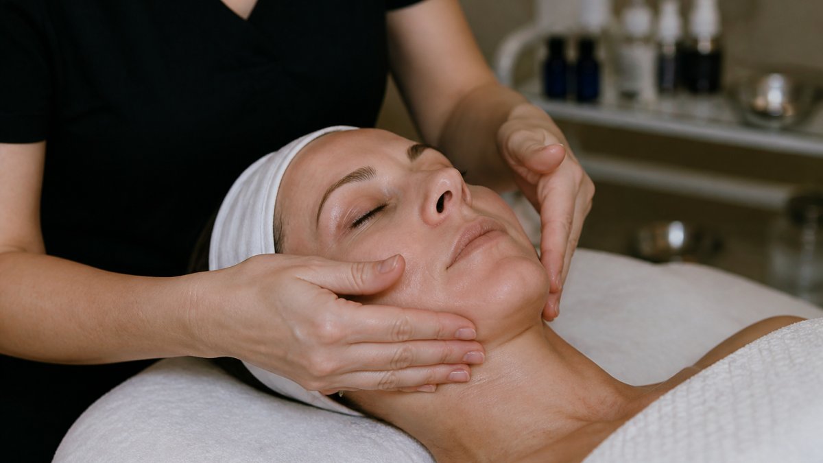 Close-up of a licensed esthetician's hands performing gentle upward sculpting strokes along a relaxed client's jawline and cheekbones in a calm, softly lit treatment room
