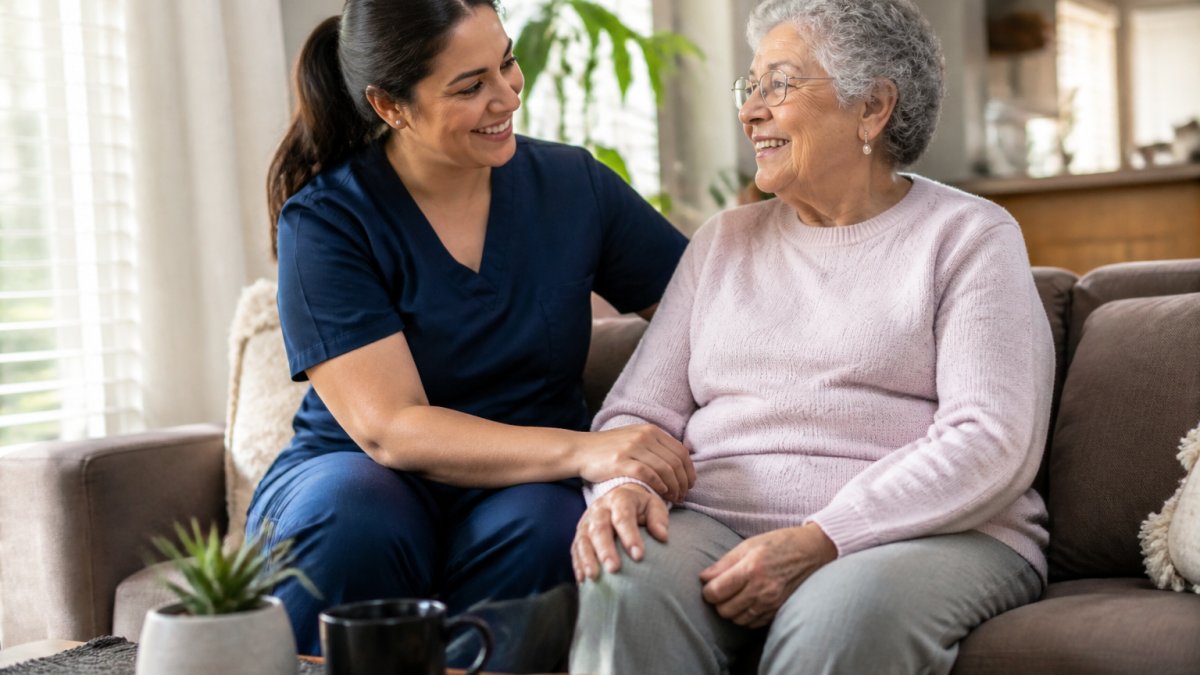 Caregiver sitting with an elderly senior at home, both smiling warmly in a bright living room with natural sunlight, showing genuine personal connection and comfort