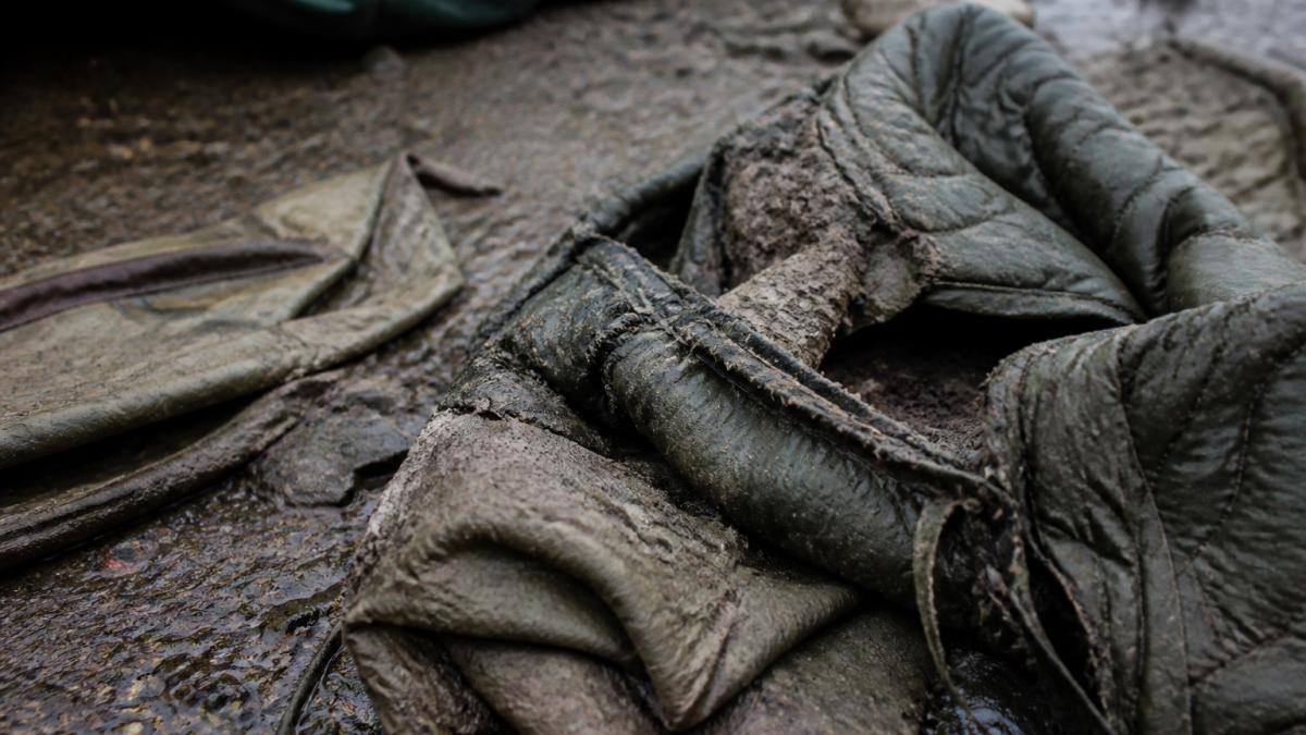 Muddy, torn outdoor gear with separated seams and worn fabric scattered on wet ground after extended field use in harsh conditions