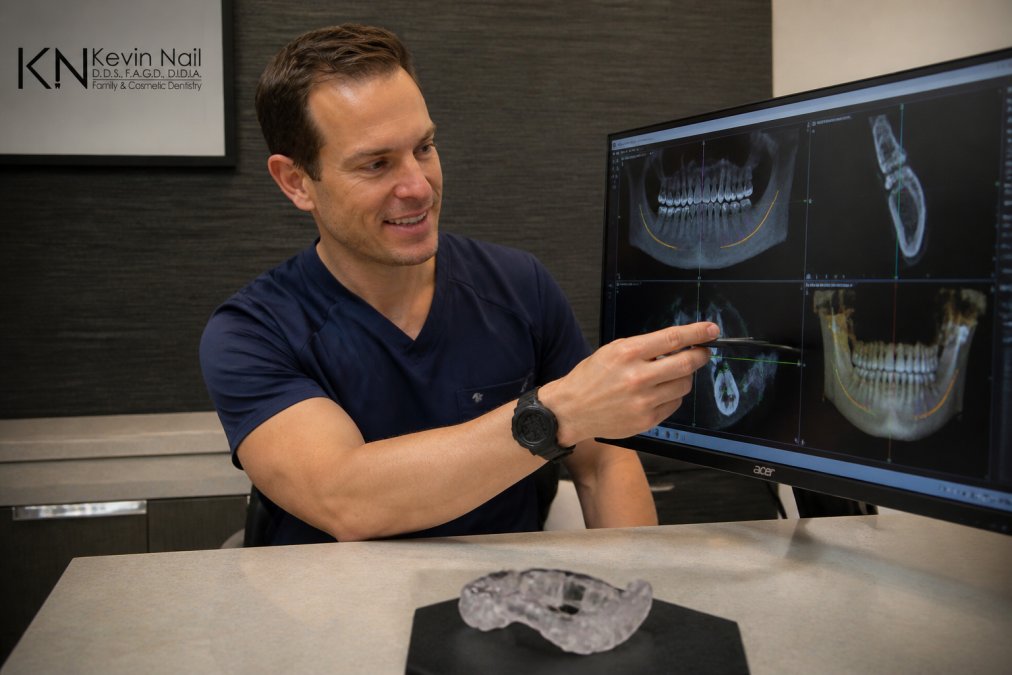 A dentist in scrubs reviewing a 3D CBCT scan on a monitor in a modern dental operatory, with implant planning software visible on the screen and a surgical guide template resting on the counter