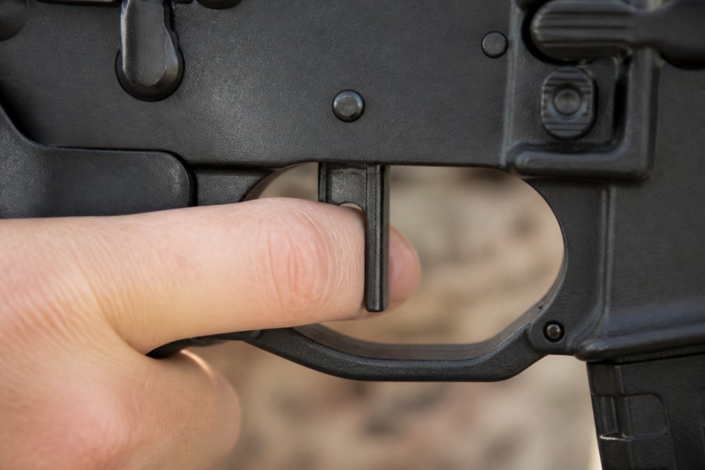 Close-up of a shooter's index finger resting on a flat-faced trigger inside an AR-15 trigger guard, showing proper finger pad contact and straight-back press alignment