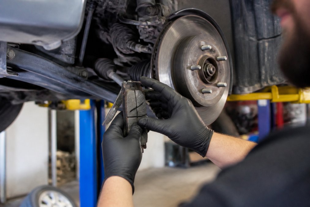 Mechanic's hands closely examining brake pads and suspension components on a vehicle lift, with diagnostic measurement tools visible and detailed wear patterns being assessed
