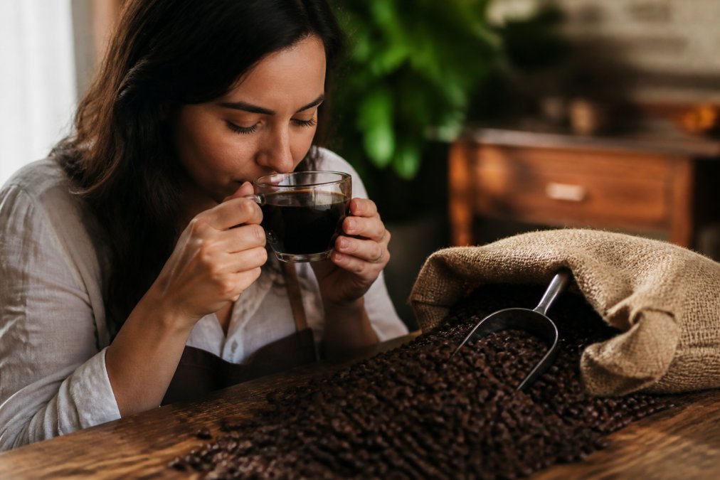 A person holding a clear glass coffee cup up to their nose, inhaling the aroma, with a burlap coffee bag and roasted beans on a wooden table nearby