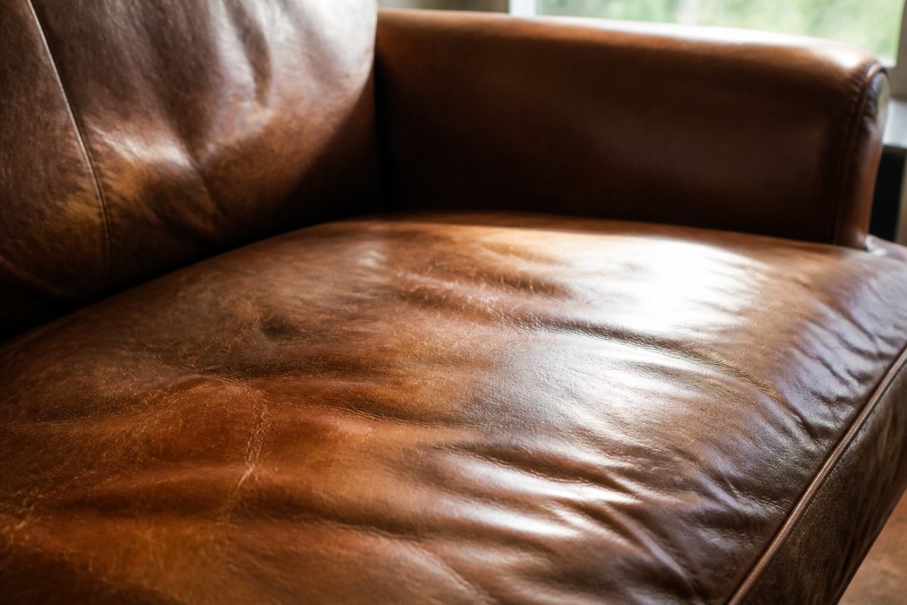 Close-up of a well-worn full grain leather sofa cushion showing natural patina, with soft window light highlighting the grain texture and subtle color variations