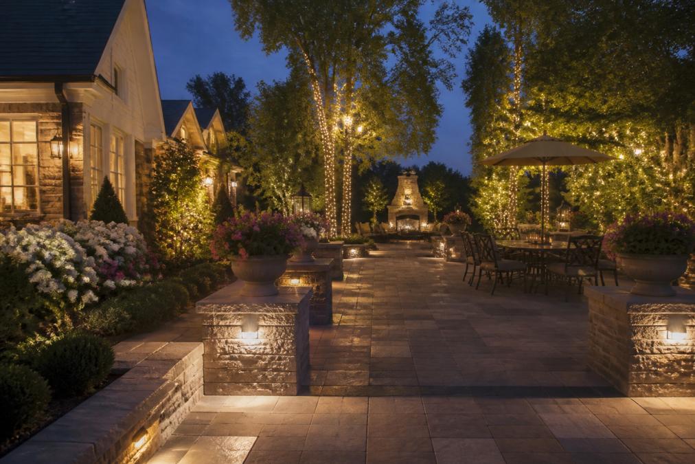 A beautifully designed upscale residential backyard at dusk, featuring a custom stone patio with ambient outdoor lighting, ornamental plantings in full bloom, and warm holiday lighting accents wrapped around mature trees and shrubs, photographed in soft golden evening light