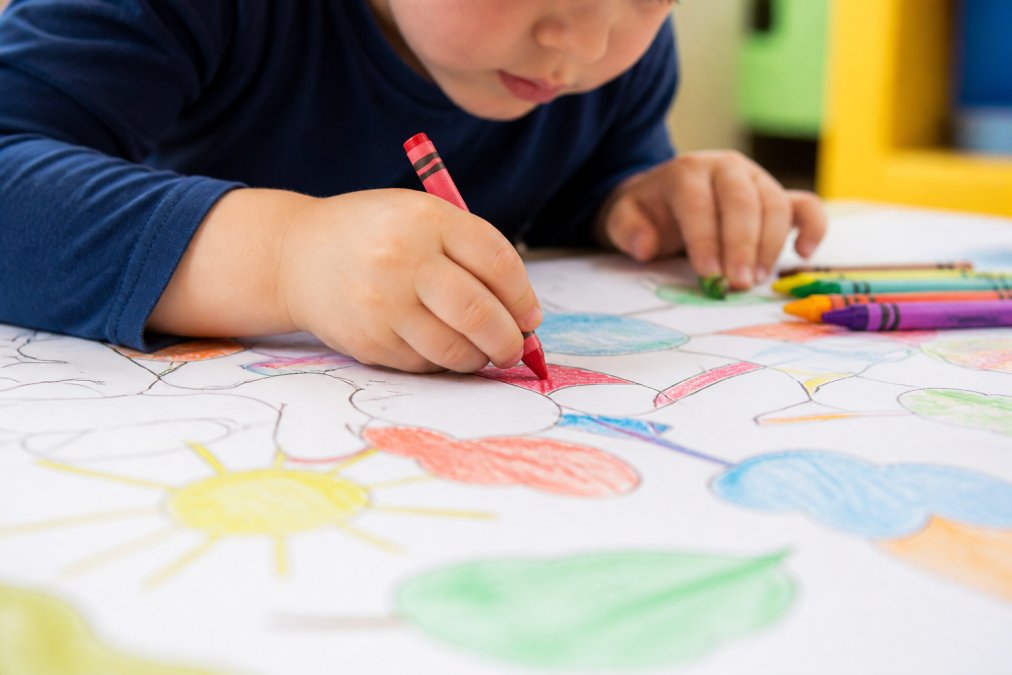 Close-up of a toddler's hand gripping a crayon over a large colorful table-sized coloring sheet, showing pencil grasp and focused concentration on filling in shapes