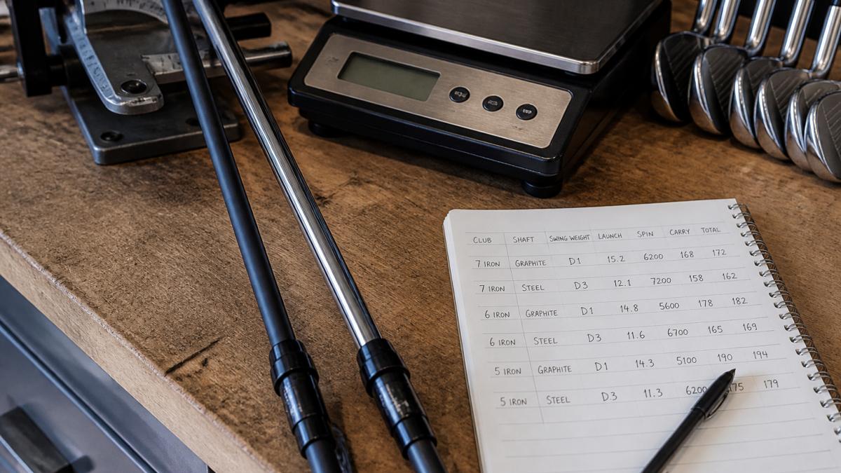 Graphite and steel golf shafts on a workbench with a swing-weight scale and launch data notebook