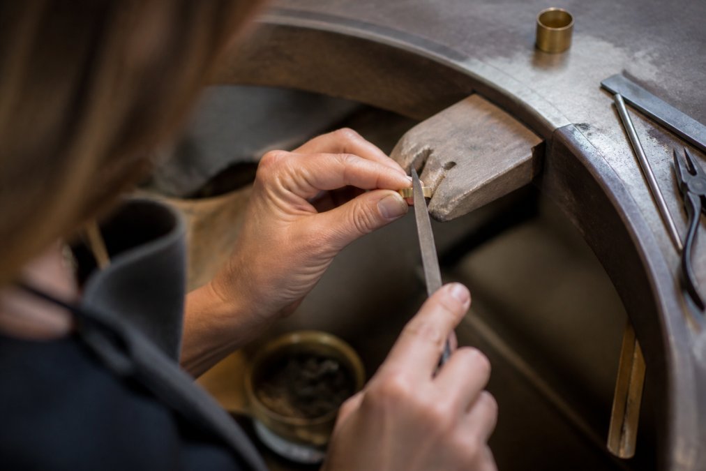 Close-up of a jeweler's hands working with metal at a jewelry bench, using hand tools to shape and fabricate a piece, with natural studio lighting