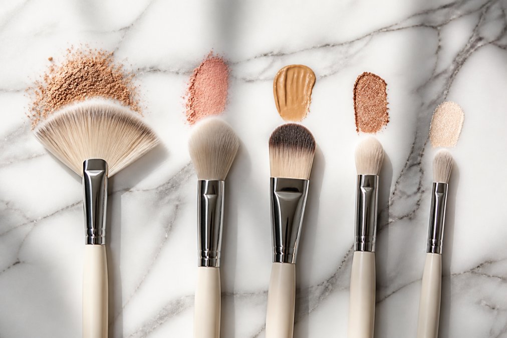 Flat lay of several makeup brushes with different head shapes, fan, tapered, flat, and dome, arranged neatly on a white marble surface alongside soft pastel makeup swatches in warm natural light