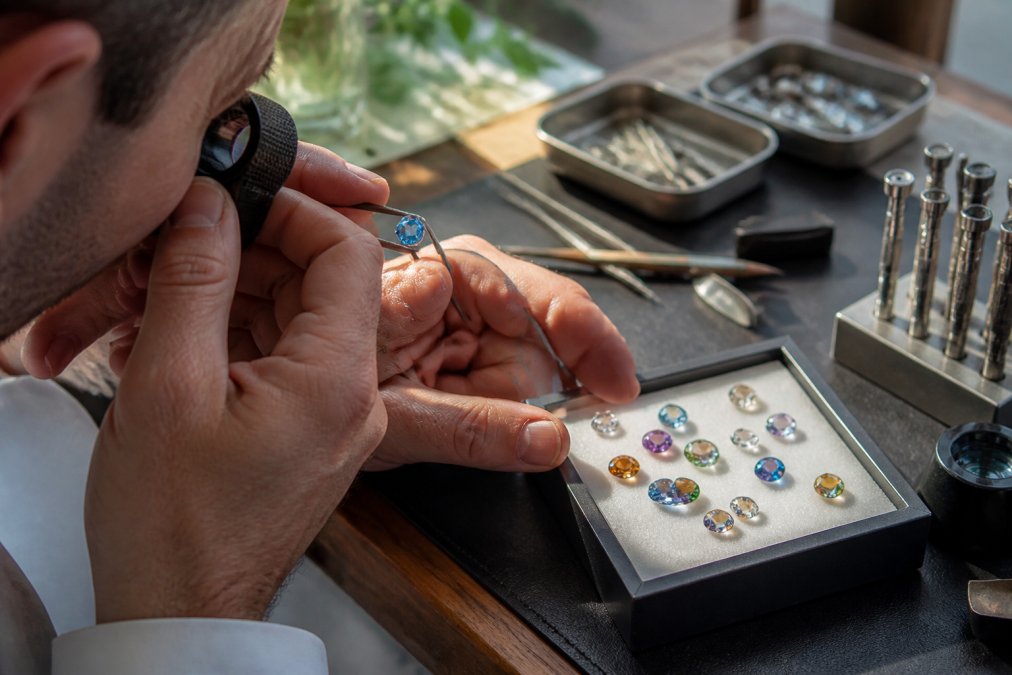 Professional jeweller's hands examining a gemstone under magnification with gemological tools, loupe, and various cut stones visible on a workbench with natural lighting