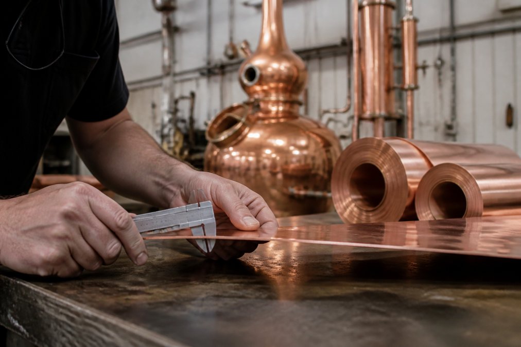 Craftsman measuring the thickness of heavy gauge copper with a caliper in a workshop with a finished still nearby