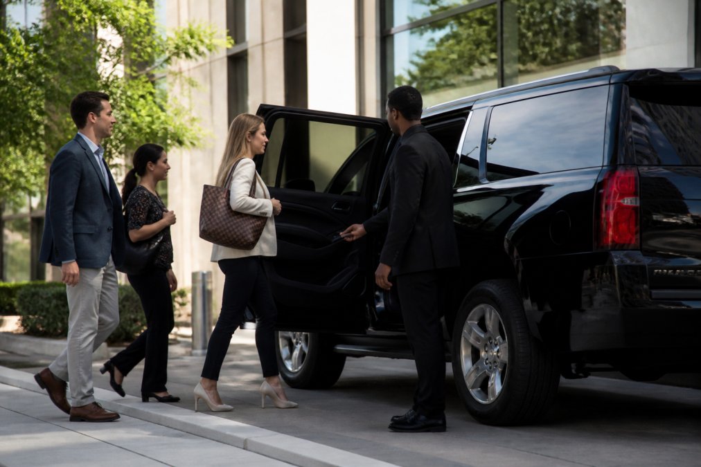 A sleek black SUV parked curbside outside a modern office building in Charlotte, with a professional chauffeur in a dark suit holding the rear passenger door open for a small group of business travelers in smart casual attire