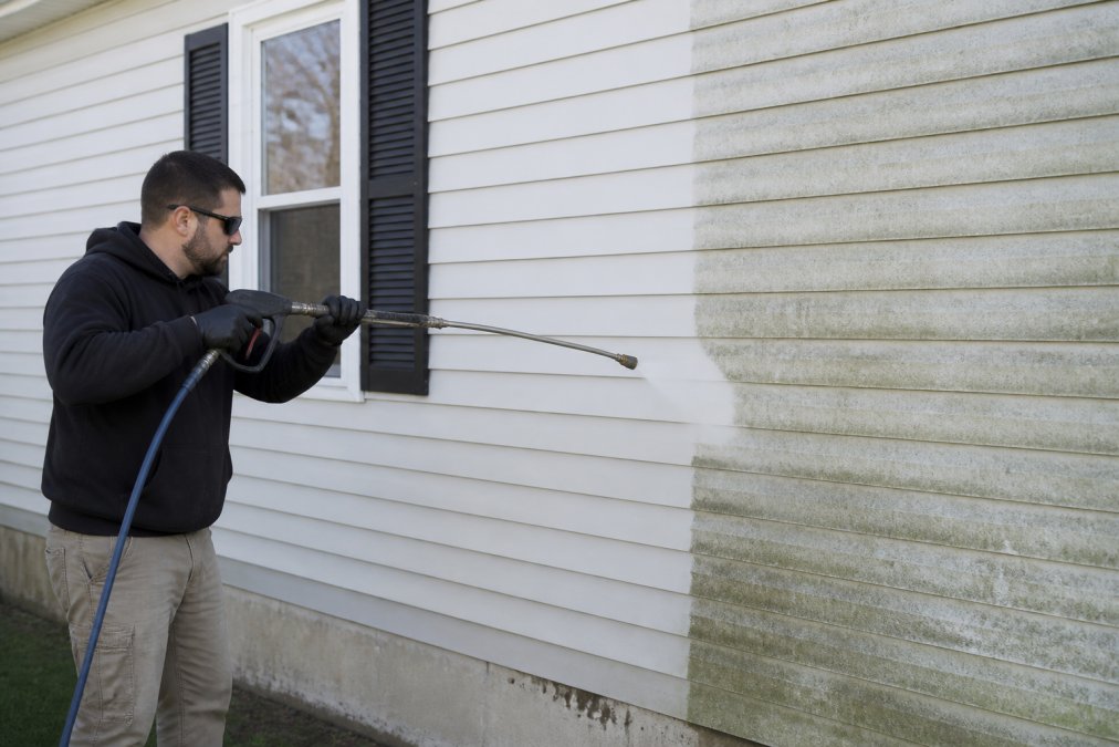 Professional exterior cleaning technician using soft-wash equipment on residential siding, with mold and algae visibly being removed, revealing clean restored surface beneath
